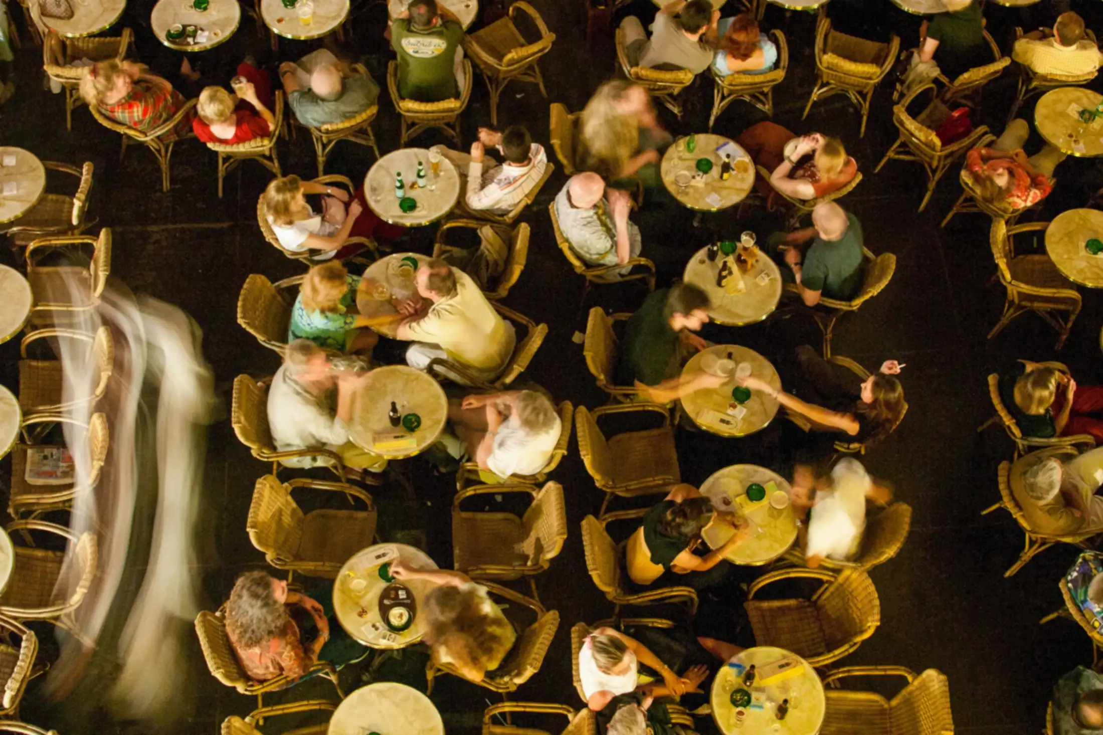 Een vol caféterras op een zaterdagavond, van bovenaf gezien, met een ober die tussen de tafels door loopt