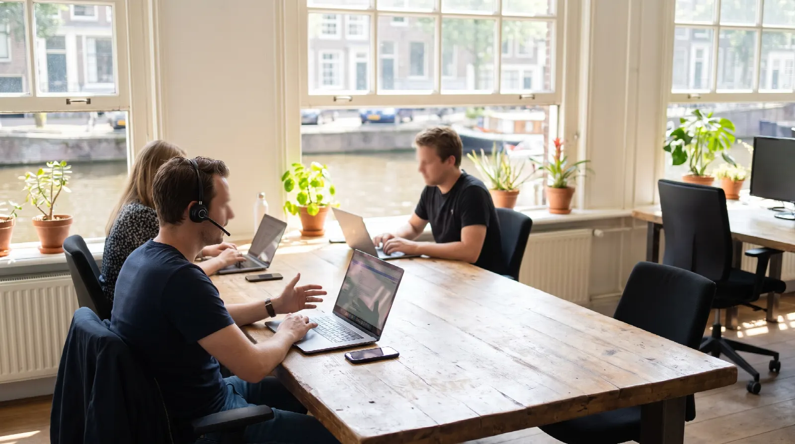 Drie collega's werken op laptops aan een houten tafel in een licht kantoor aan de gracht, een van hen belt met een headset